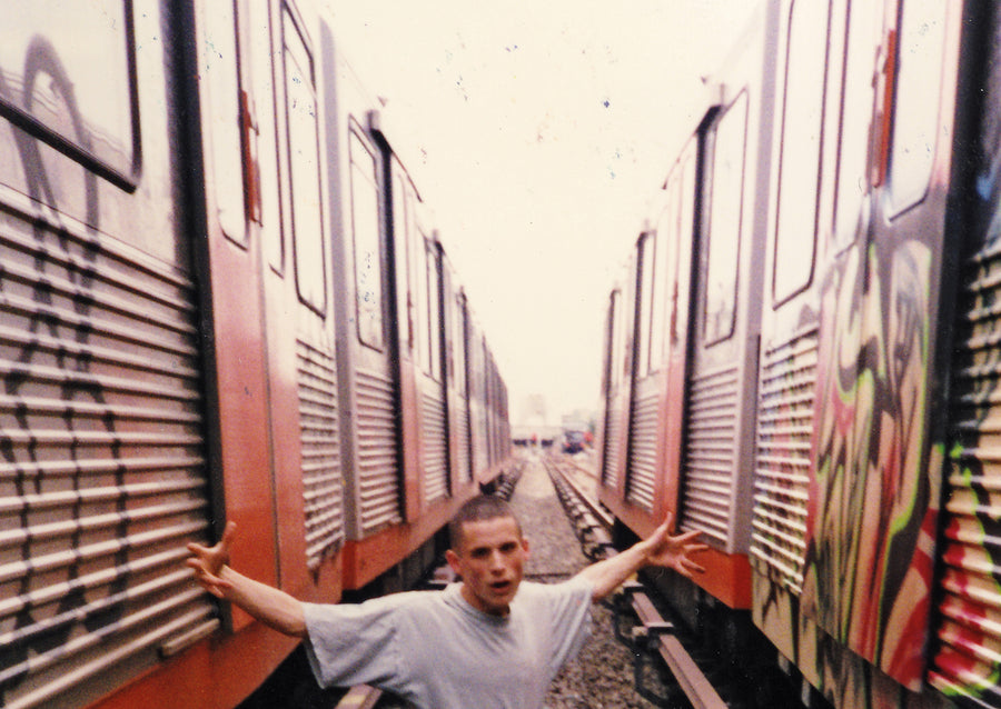 Writer with outstretched arms between two graffiti-covered trains in a yard