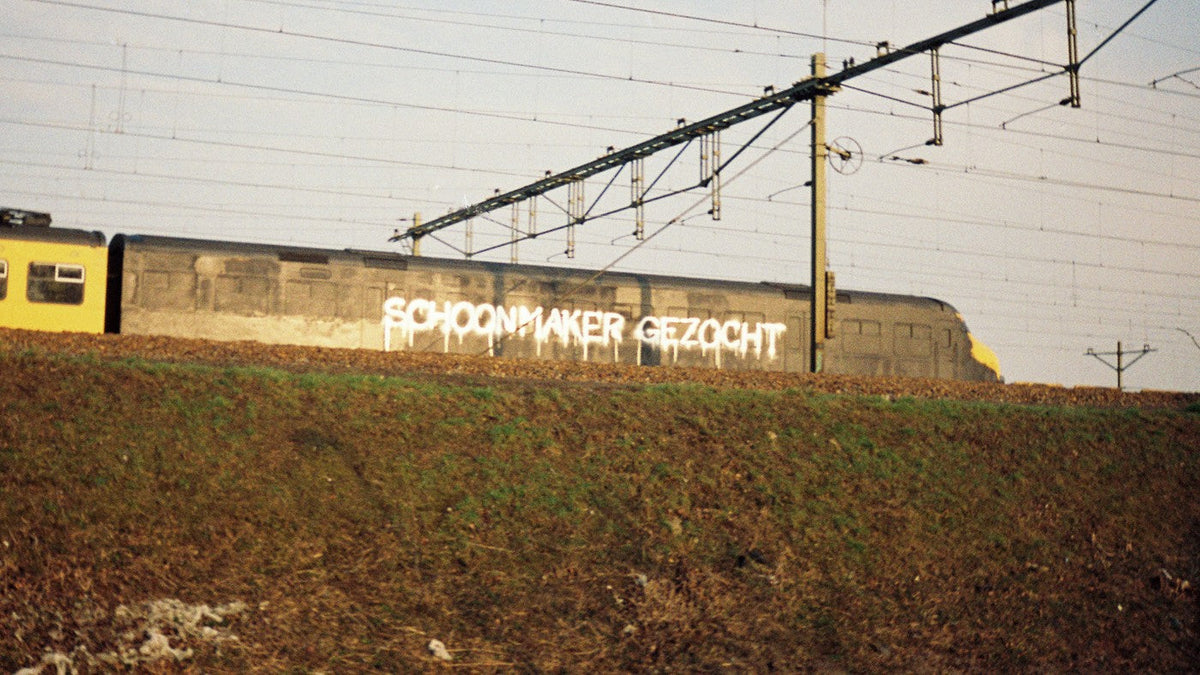 Grey Dutch train with the words 'Schoonmaker gezocht' sprayed in dripping white letters, seen from below an embankment