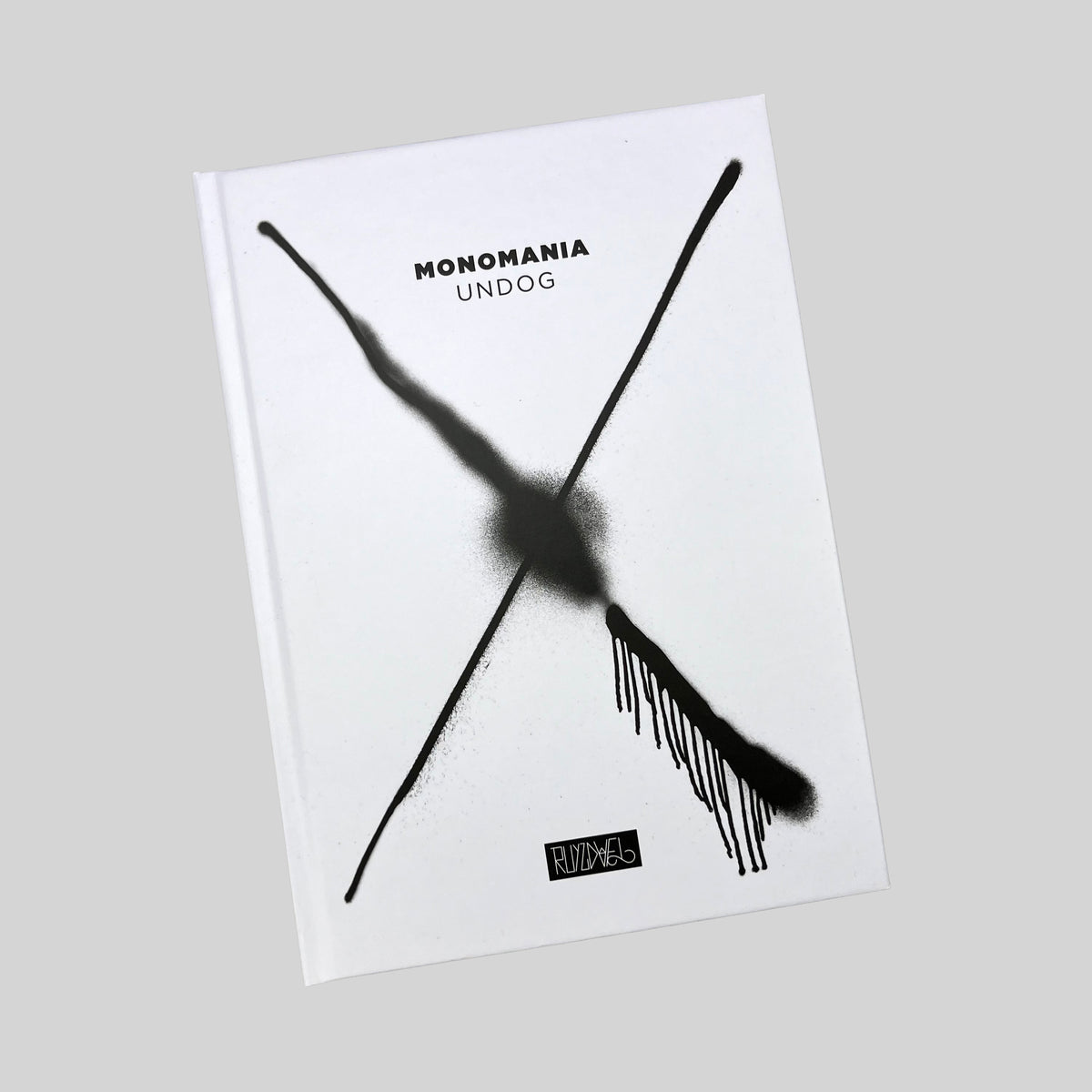 MONOMANIA, white hardcover with a black spray-painted cross, seen at an angle