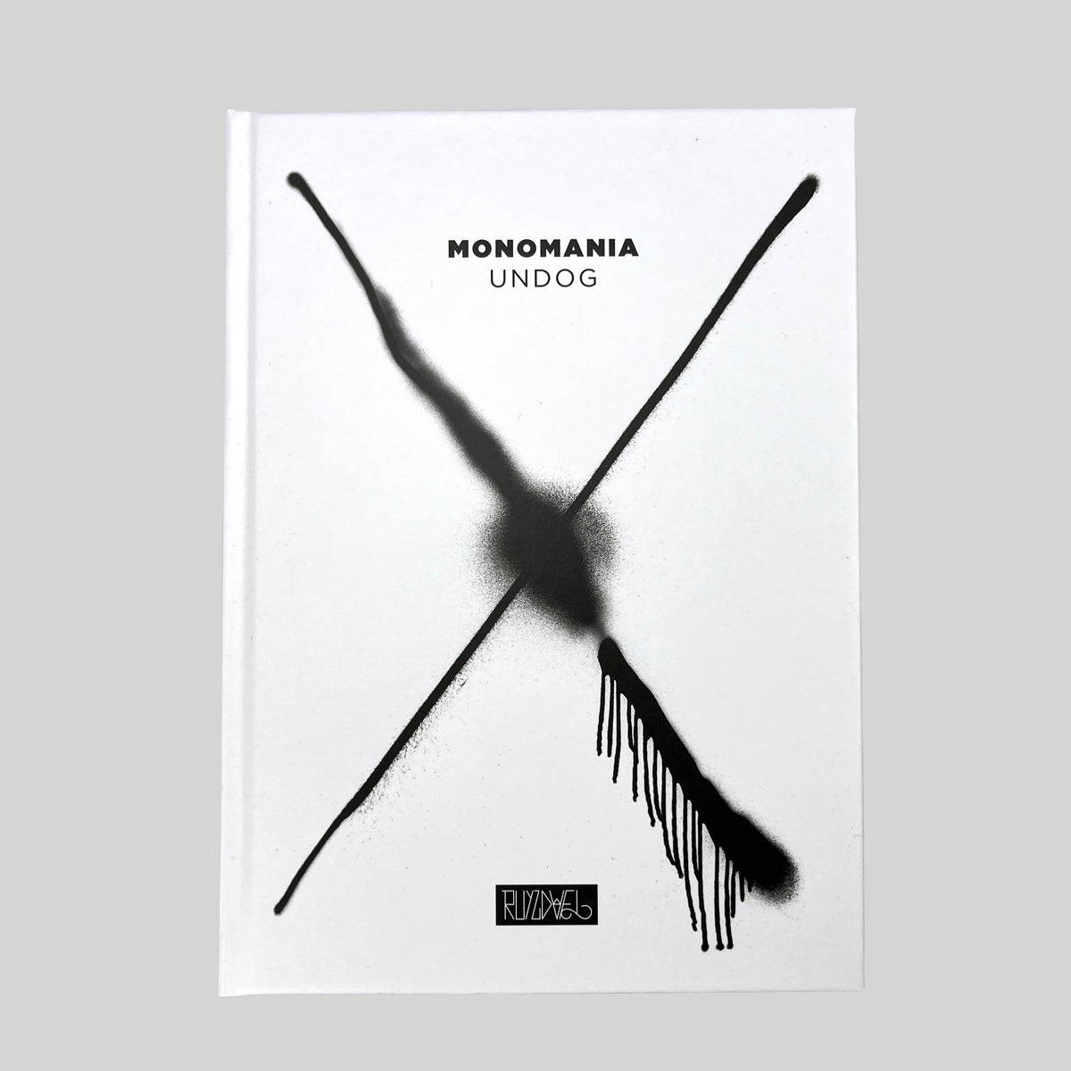 MONOMANIA, front of the white hardcover with a black spray-painted cross